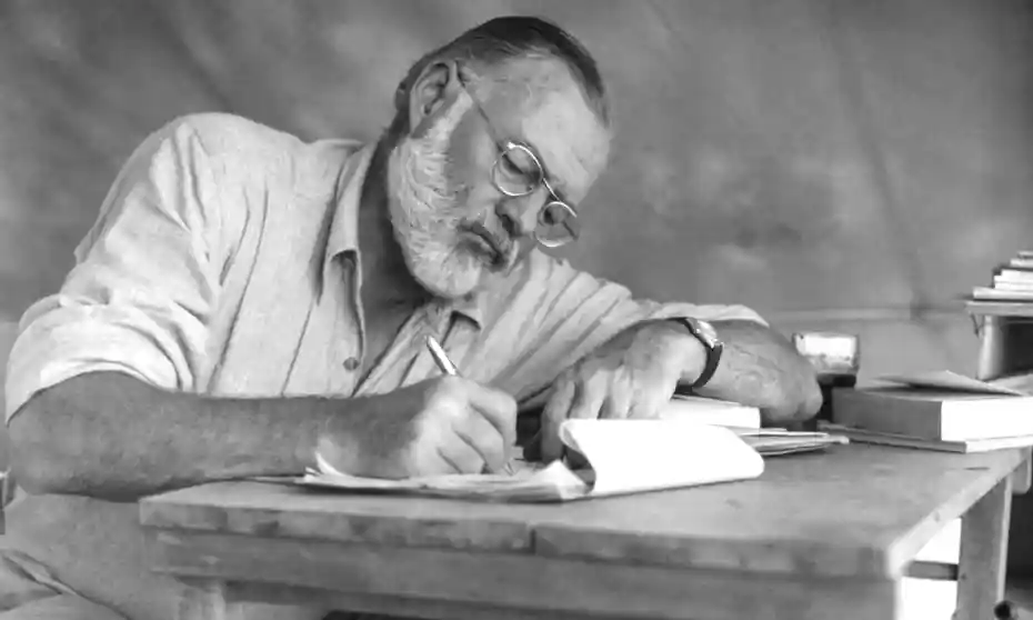 Ernest Hemingway writing at desk with pen and paper.